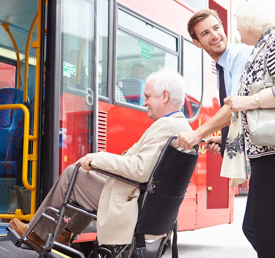 An elderly man in a wheelchair is being assisted onto a red bus by a young man, while a woman stands nearby.