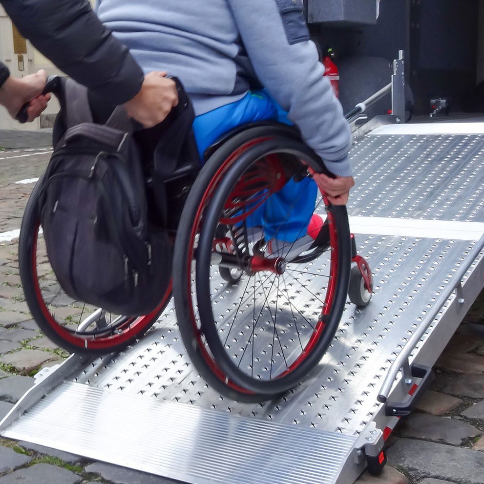 A person assists a wheelchair user onto a van using a metal ramp on a cobblestone street.